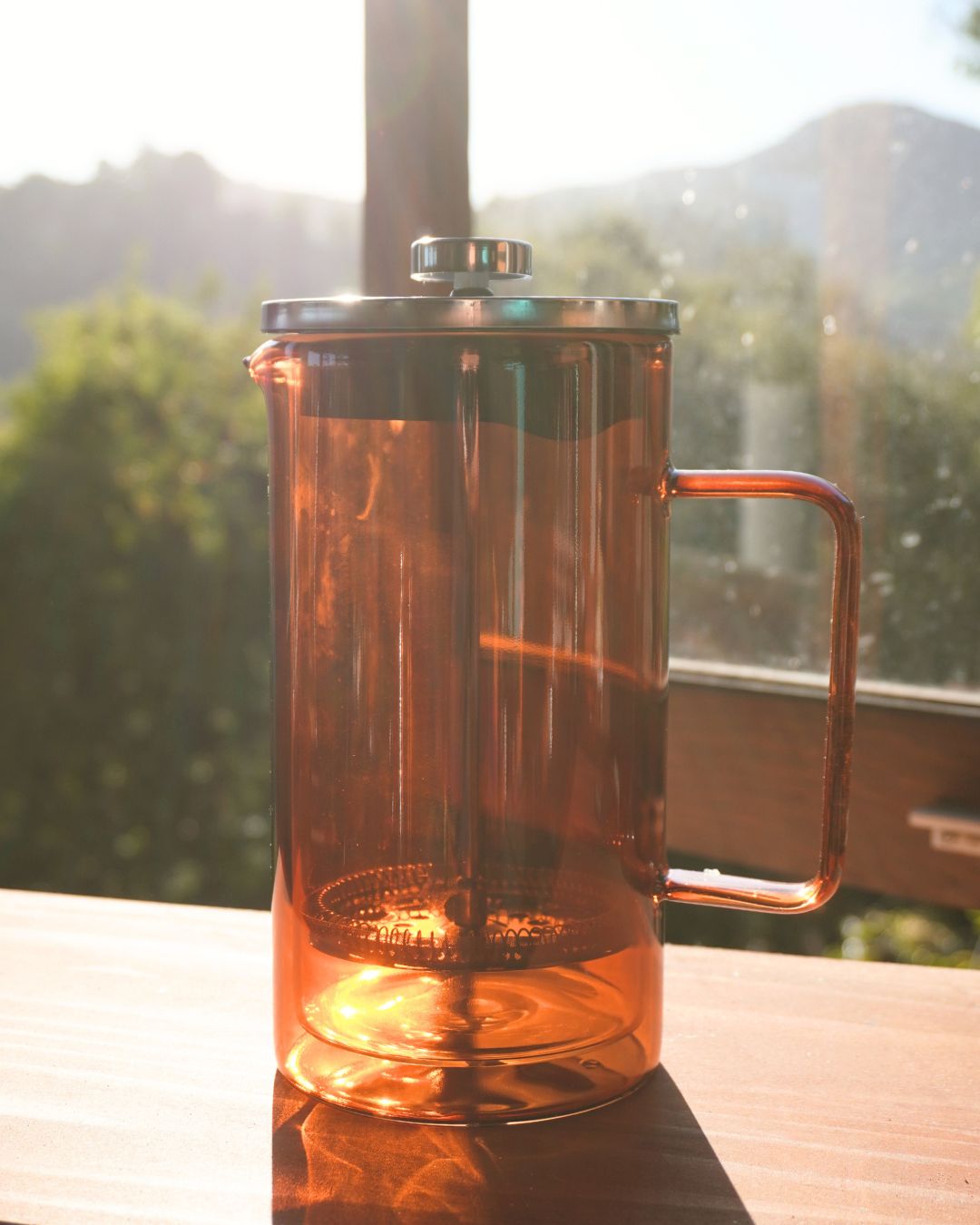 Glass French press coffee maker with stainless steel plunger on a sunlit kitchen table