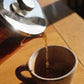 Fresh coffee pouring from a glass French press into a ceramic mug